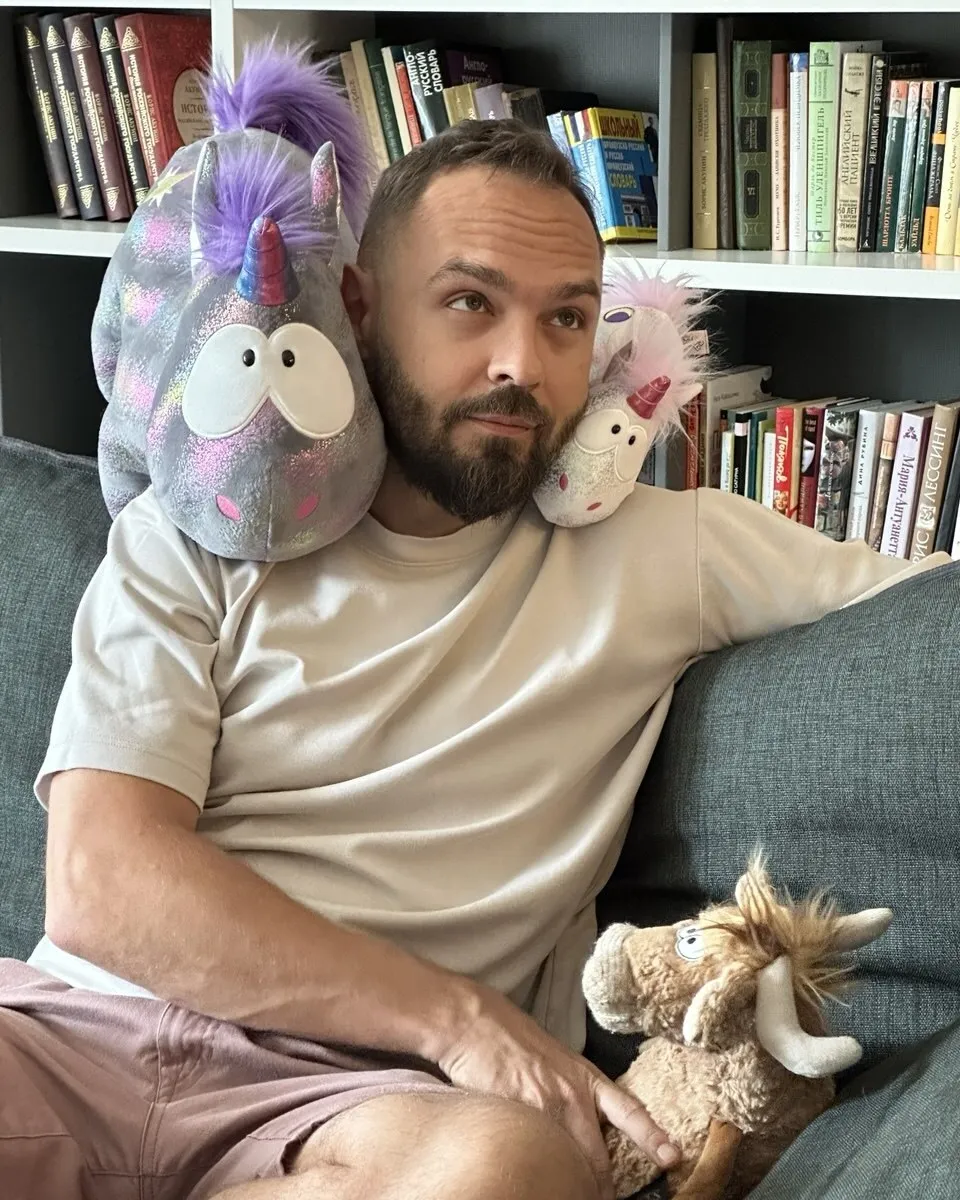 Andrey on a sofa in front of bookshelves, with two plush unicorns perched on the shoulders and a plush bull alongside